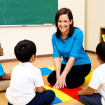 A professional female kindergarten teacher, fully clothed in a modest, comfortable professional top and trousers, is kneeling gracefully to interact with a diverse group of 4-5 young children (boys and girls, 4-5 years old) in a brightly lit, inviting kindergarten classroom. She is smiling genuinely, looking directly at the children, who are happily engaged in a creative activity like building with blocks or drawing. The classroom is filled with colorful, age-appropriate learning materials and comfortable child-sized furniture. The scene captures a moment of nurturing guidance and joyful learning, emphasizing the teacher's positive impact on child development. Soft, natural lighting. Professional photography, high resolution, perfect anatomy, correct proportions, natural pose, well-formed hands, proper finger count, natural body proportions, safe for work, appropriate content, fully clothed, modest, family-friendly.